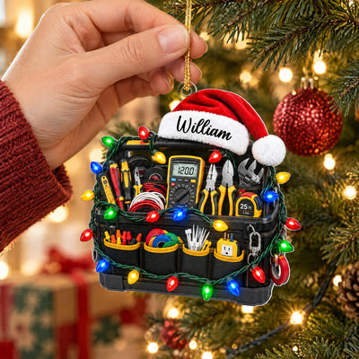 Personalized electrician tool bag acrylic ornament held by a hand in a red sleeve near a decorated tree