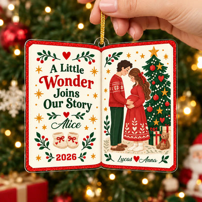 Personalized acrylic book ornament held by hand in front of a lit Christmas tree with red baubles