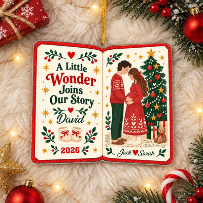 Personalized acrylic book ornament held by hand in front of a lit Christmas tree with red baubles