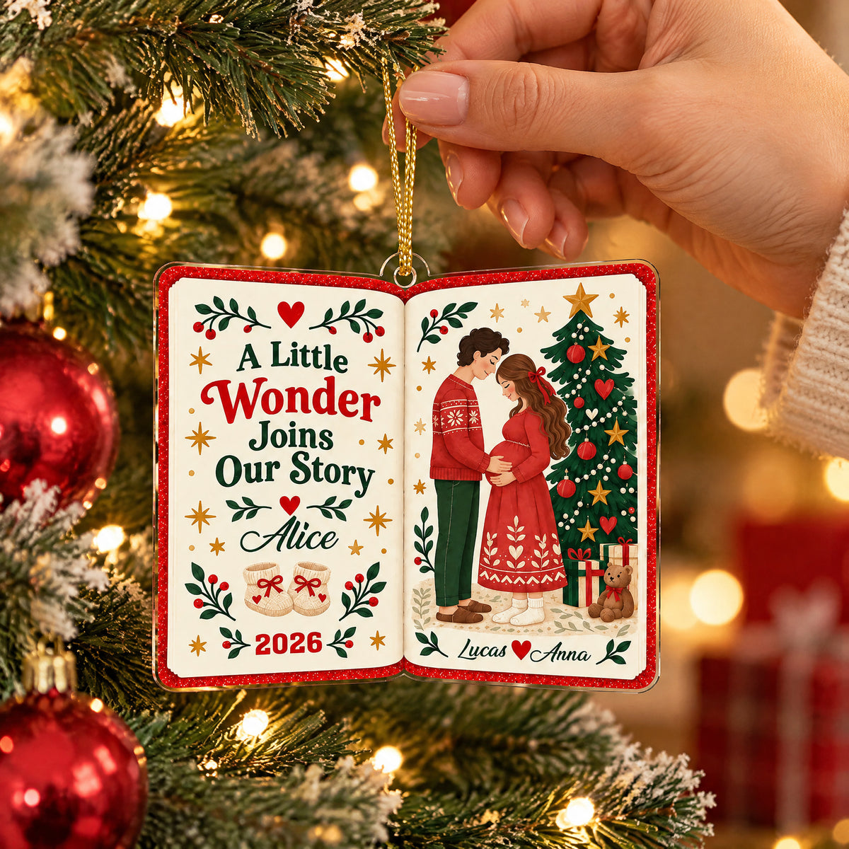 Personalized acrylic book ornament held by hand over a snowy tree branch, red baubles at left