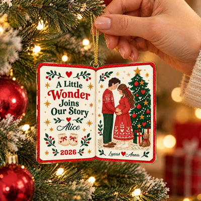 Personalized acrylic book ornament held by hand over a snowy tree branch, red baubles at left