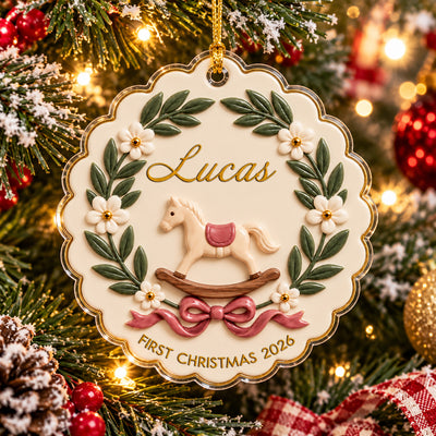 Personalized rocking horse baby acrylic ornament hanging on a snowy tree beside red baubles and plaid ribbon