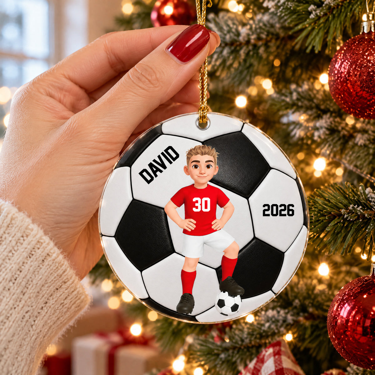 Personalized soccer acrylic ornament held in a hand with red nails beside a lit Christmas tree and red baubles