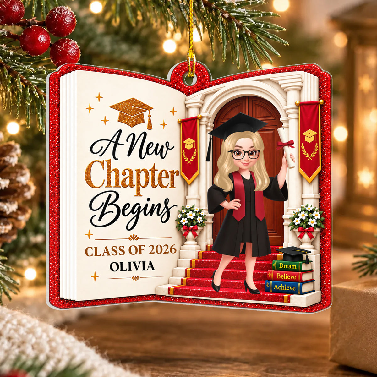 Personalized acrylic graduate ornament shaped like an open book, hanging on a pine branch with warm lights and a pinecone