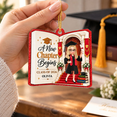 Personalized acrylic graduate ornament held by a hand beside a graduation cap with a gold tassel on a wooden table