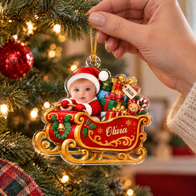 Personalized acrylic ornament baby sleigh held by a hand on its gold cord beside a lit tree