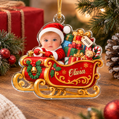 Personalized acrylic ornament baby sleigh standing on a wooden table with gift box, pinecone and red baubles