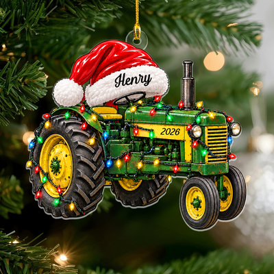 Personalized tractor acrylic ornament with Santa hat reading Henry, hanging on a pine branch with blurred lights
