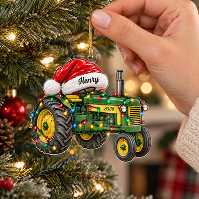 Personalized tractor acrylic ornament hanging from a gold cord on a lit tree, held by a hand in a knit sleeve