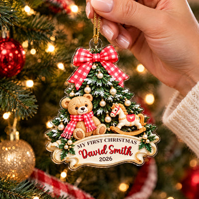 Personalized Teddy Bear Acrylic Ornament with red bow held by a hand beside lights and a gold bauble