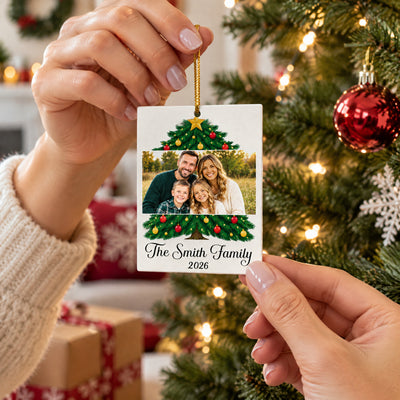 Personalized family photo name acrylic ornament held by hand near a lit tree with wrapped gifts in the background