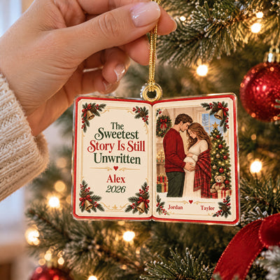 Personalized acrylic book ornament held by a hand by its gold cord in front of a lit Christmas tree