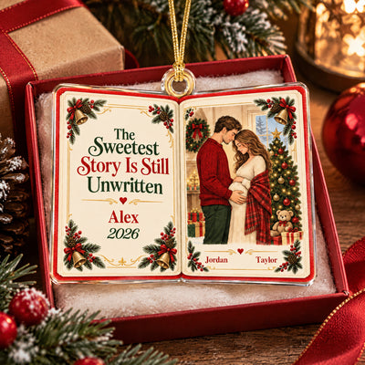 Personalized acrylic book ornament resting in a red gift box on white filling with pinecones and ribbon nearby