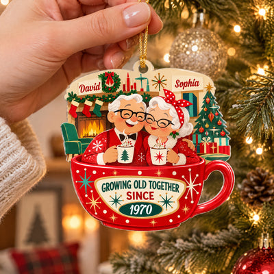 Personalized Christmas acrylic ornament held by a hand on a gold cord, with a lit tree and pine cone in the background