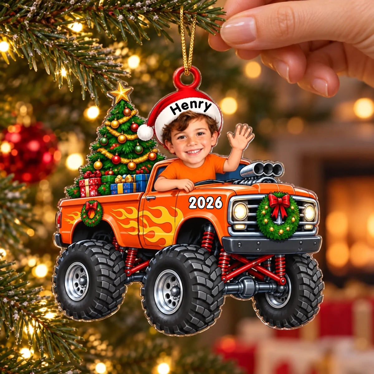 Personalized orange monster truck acrylic ornament held by a hand in front of a lit Christmas tree