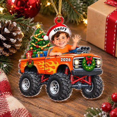 Personalized orange monster truck acrylic ornament lying on a wooden table with a wrapped gift and pinecone