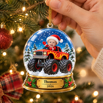 Personalized monster truck snow globe acrylic ornament held by a hand beside tree lights and a plaid bow