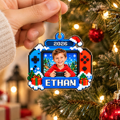 Personalized Pixel Game Console Acrylic Ornament dangling from a gold cord in a hand, tree and red ornament behind