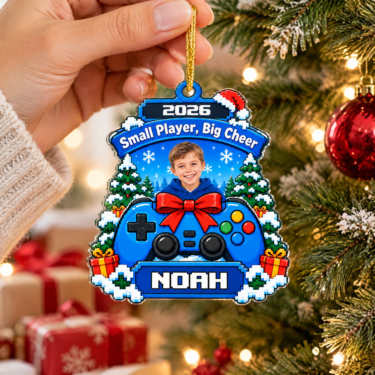 Personalized acrylic gamer ornament held by a hand on a gold cord, with wrapped gifts blurred behind