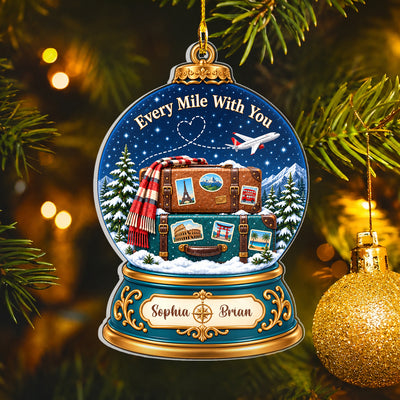 Personalized acrylic snow globe ornament beside a gold glitter bauble on a tree with blurred lights