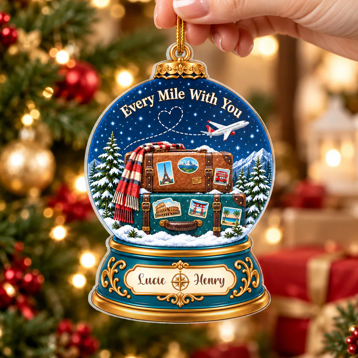 Personalized acrylic snow globe ornament held by a hand by its gold cord in front of a decorated tree and gifts