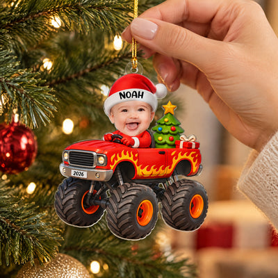 Personalized red monster truck acrylic ornament held by a hand beside a lit Christmas tree and red bauble