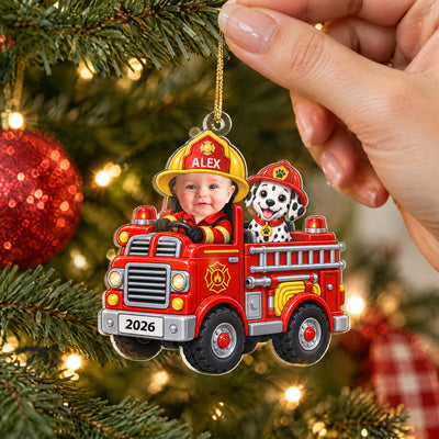 Personalized acrylic ornament baby fire engine held by a hand near a red bauble and tree lights, name ALEX