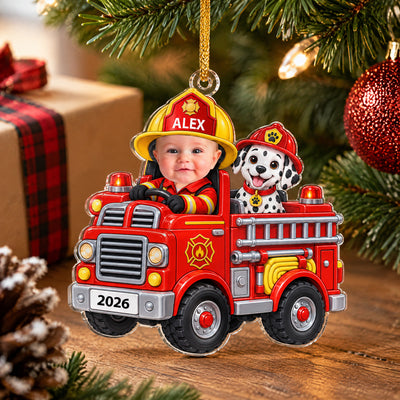 Personalized acrylic ornament baby fire engine on a wooden surface beside a plaid wrapped gift and pinecone