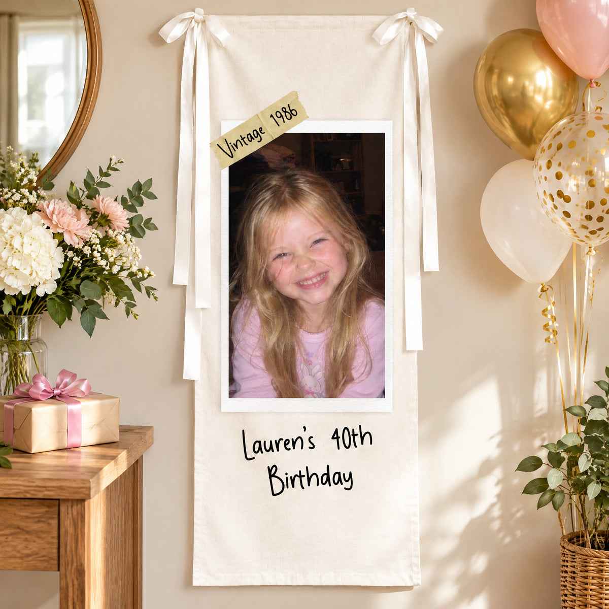 Personalized vertical photo party backdrop on a wall beside a flower vase, wrapped gift and gold balloons