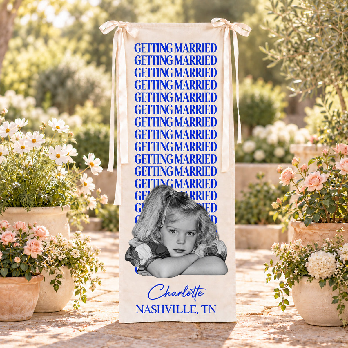 Personalized bachelorette party backdrop banner in blue text standing among flower pots in a sunny garden