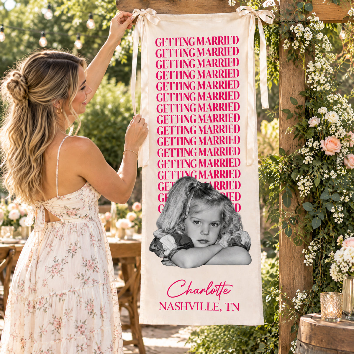 Personalized bachelorette party backdrop banner in pink being hung by a woman on a flower-covered wooden frame