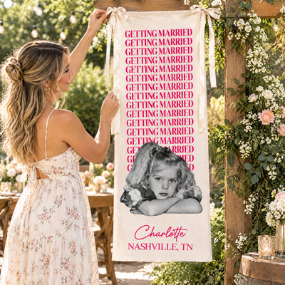 Personalized bachelorette party backdrop banner in pink being hung by a woman on a flower-covered wooden frame