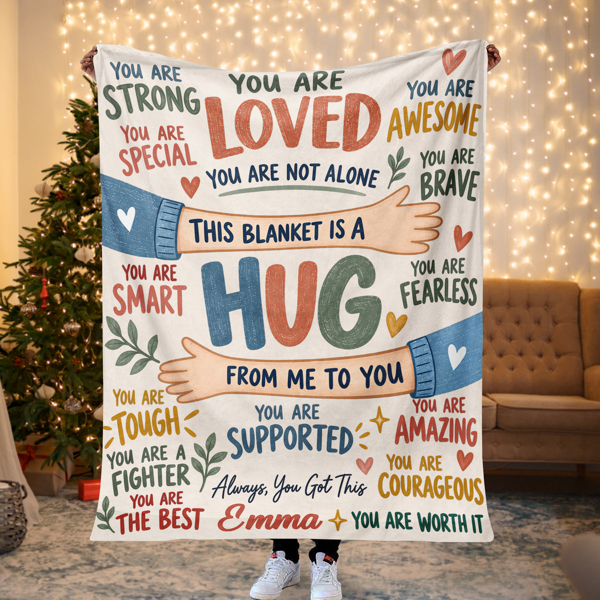 Personalized healing hug fleece blanket held up by a lit Christmas tree and string lights, name Emma shown