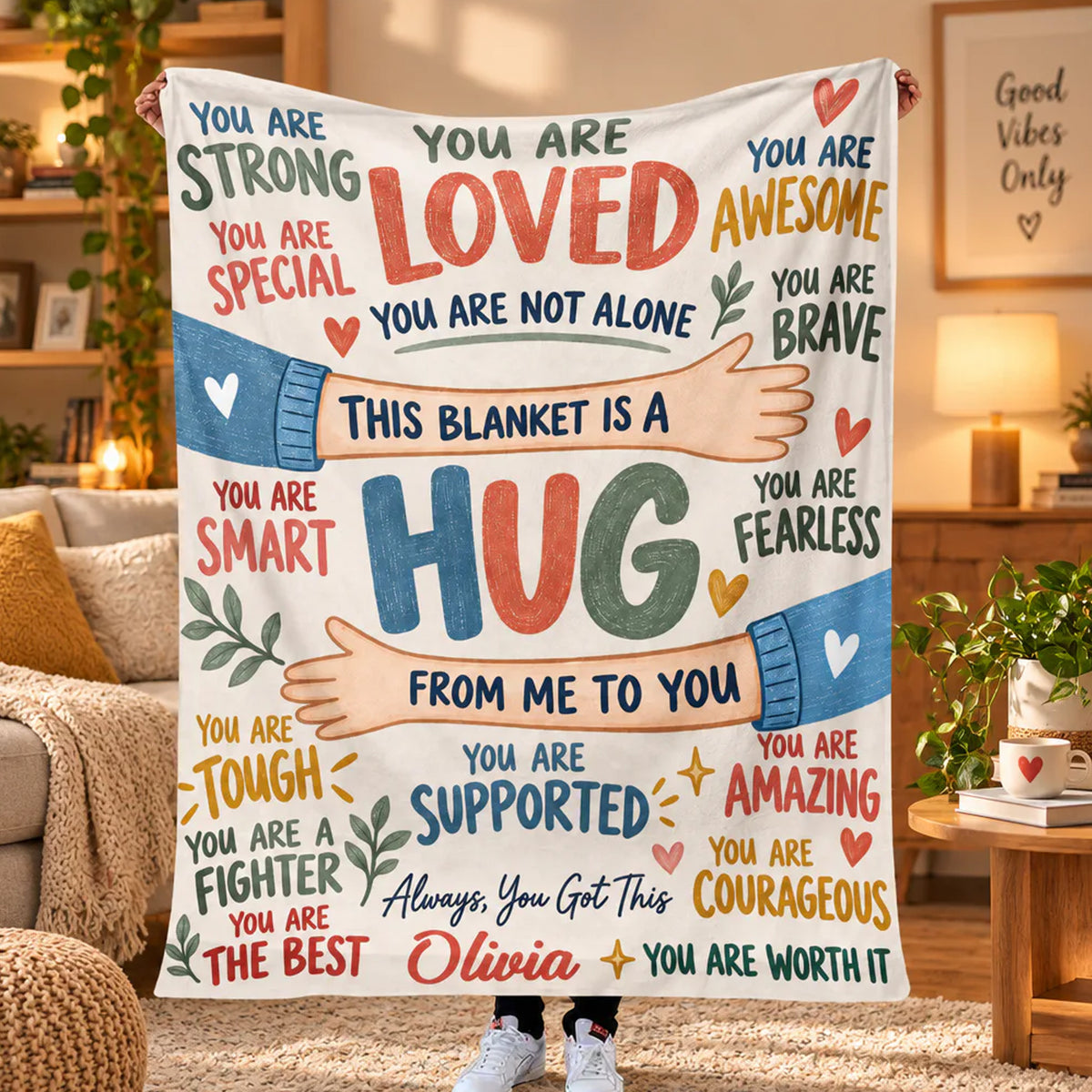 Personalized healing hug fleece blanket held up in a cozy living room with plants, name Olivia at bottom
