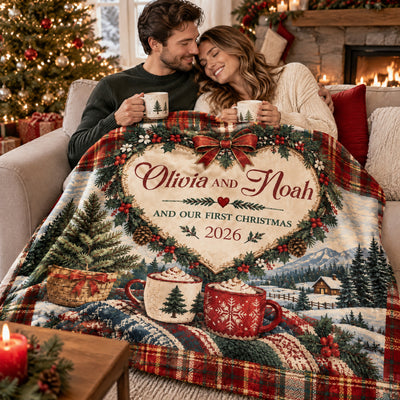 Our First Christmas Personalized Blanket covering a smiling couple holding mugs on a sofa by the fireplace