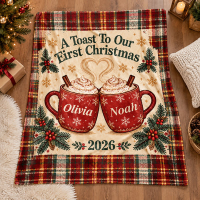 Personalized First Christmas Blanket laid flat on a wood floor with a gift, candle lantern and knit pillow nearby
