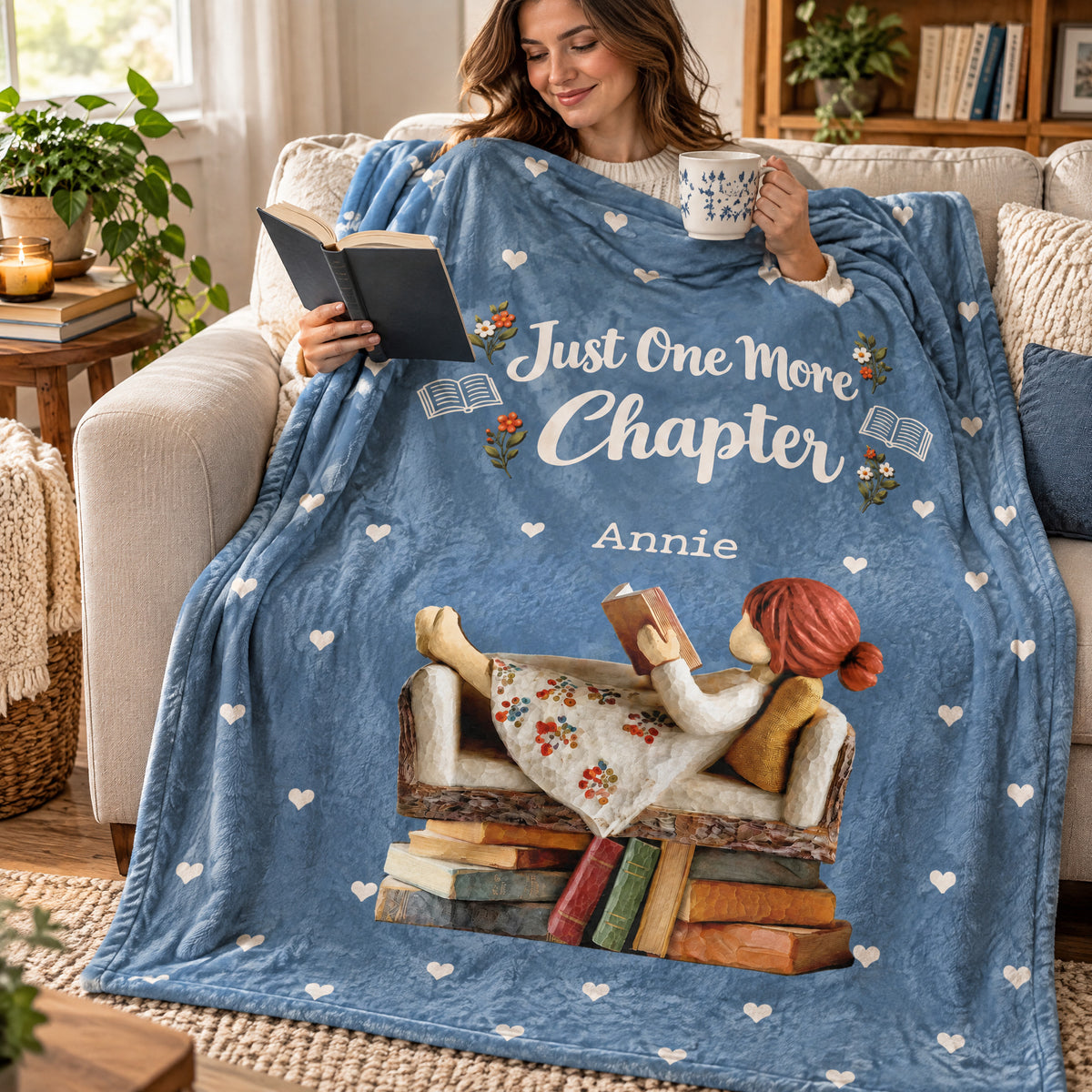 Personalized Blanket in blue wrapped around a smiling woman reading a book and holding a mug on a sofa