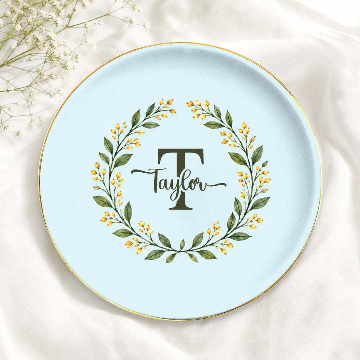 Monogram botanical wreath custom ceramic dish in light blue with Taylor name, gold rim, on white fabric beside baby's breath