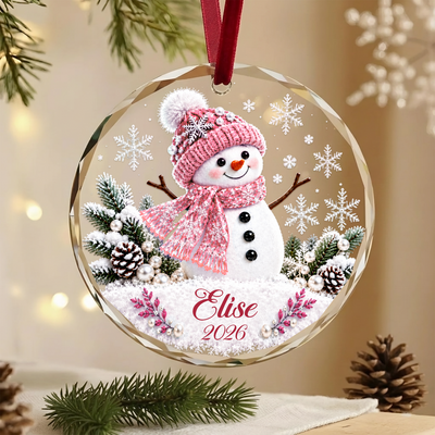 Personalized snowman glass ornament with pink hat and scarf, name Elise 2026, hanging by a red ribbon near pine