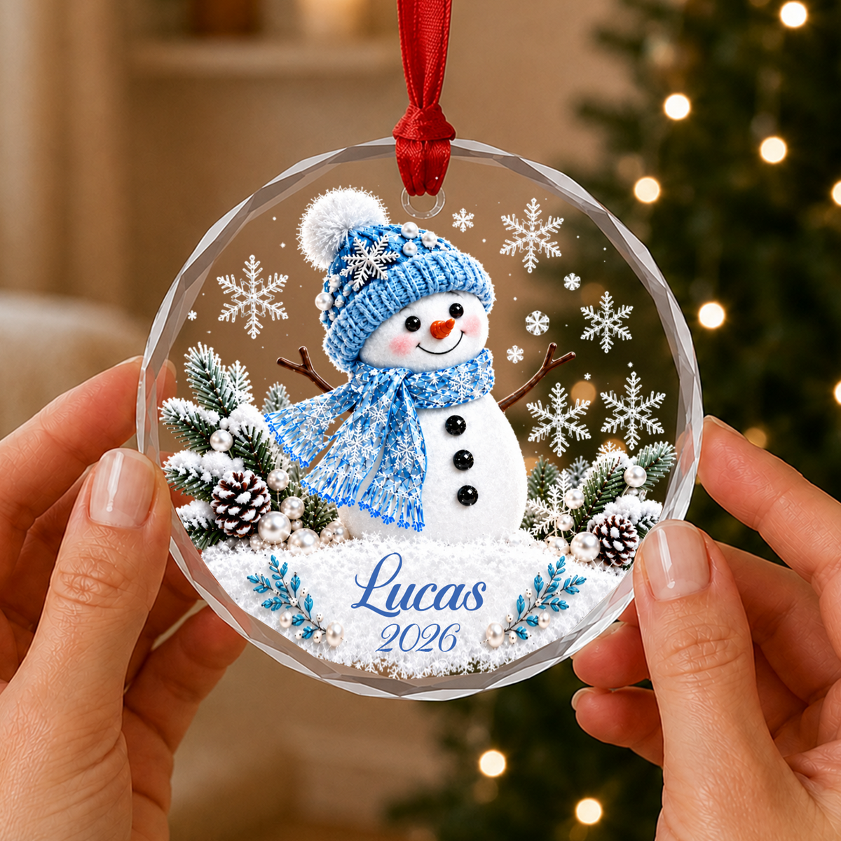 Personalized snowman glass ornament with blue hat and scarf, name Lucas 2026, held in two hands by a lit tree