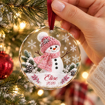 Personalized snowman glass ornament with pink scarf held in one hand against tree branches and a red bauble