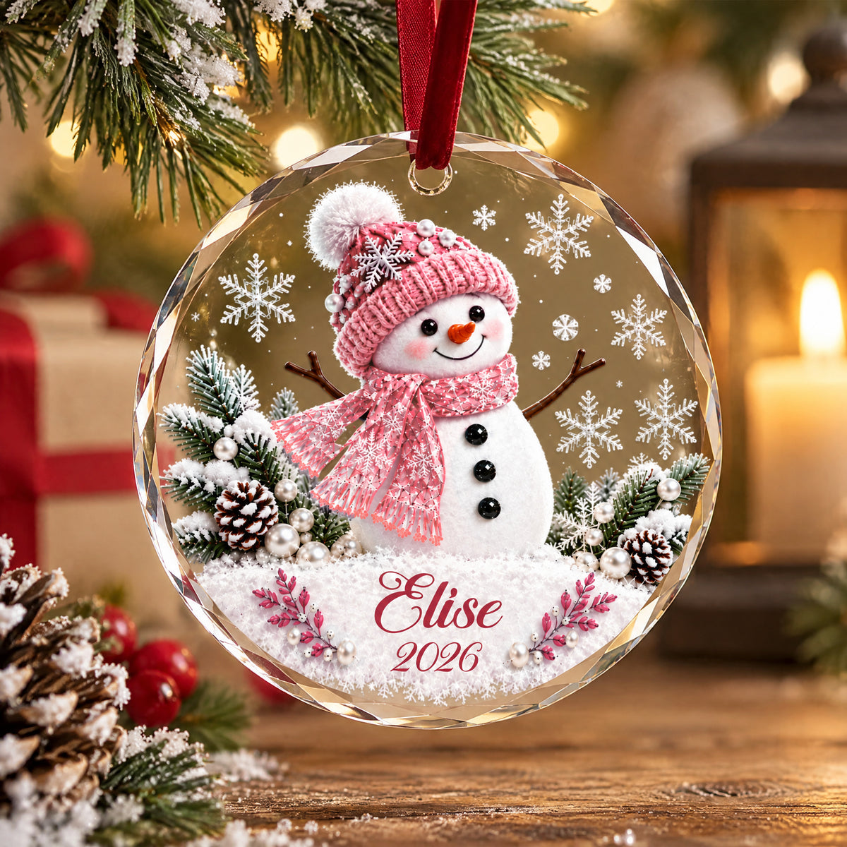 Personalized snowman glass ornament with pink hat hanging on a branch, candle lantern and pinecones on a wooden table