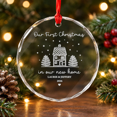Personalized glass ornament new home with red ribbon, beside pine branches, a pinecone and warm lights