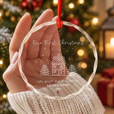 Personalized glass ornament new home with red ribbon, beside pine branches, a pinecone and warm lights