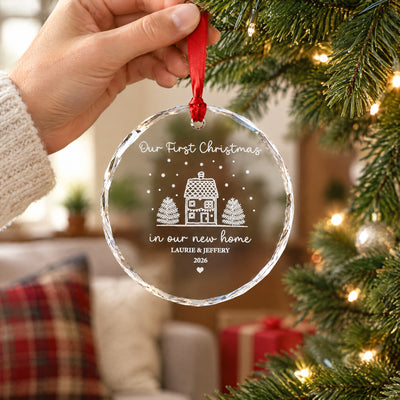 Personalized glass ornament new home held by red ribbon in a hand, in front of a tree, plaid pillow and gift