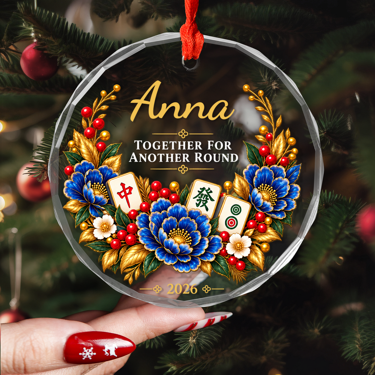 Personalized glass ornament with mahjong held in a hand near tree branches, showing the name Anna and 2026