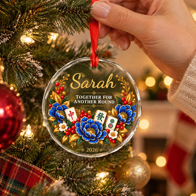 Personalized glass ornament with mahjong held by a red ribbon on a lit tree with a stocking in the background