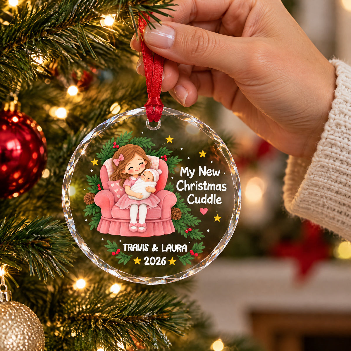 Personalized glass ornament My New Christmas Cuddle held by a hand in a knit sleeve in front of a tree