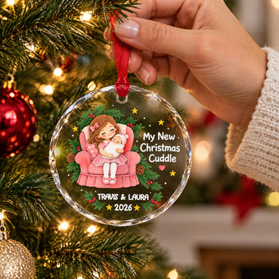 Personalized glass ornament My New Christmas Cuddle held by a hand in a knit sleeve in front of a tree