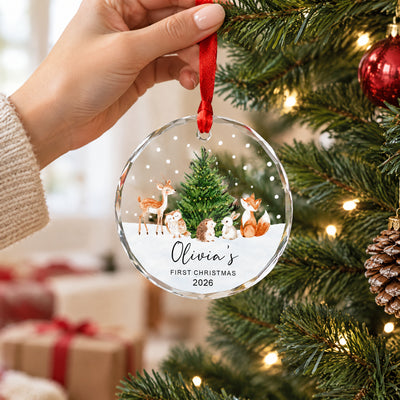 Personalized Woodland Animals Glass Ornament held by a hand beside tree branches, a pinecone and wrapped gifts
